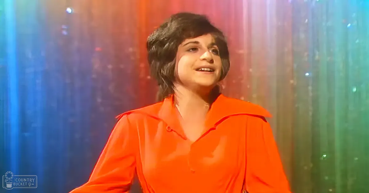A young adult female wearing a relaxed, long-sleeved, collared, bright orange shirt with buttons visible down the front stands against a colorful backdrop with vertical stripes of red, blue, green, and other colors. The setting appears to be indoors during evening or night due to artificial lighting.