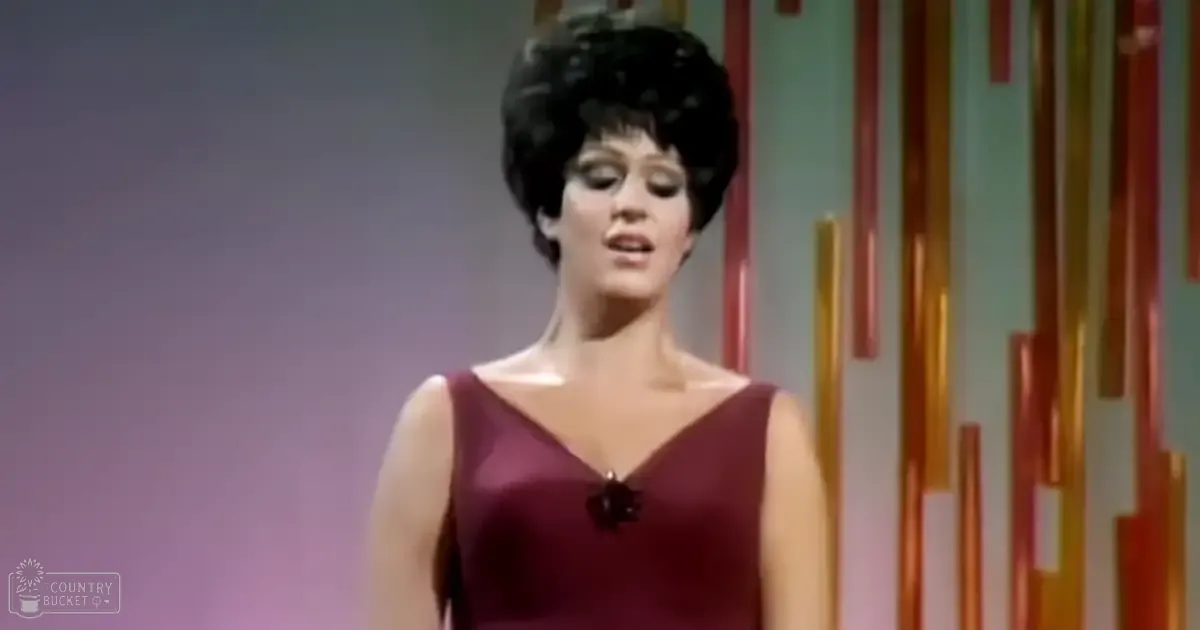 An adult female performer in a sleeveless maroon dress with a deep V-neckline, performing on stage with vertical orange and red stripes as part of the set design.