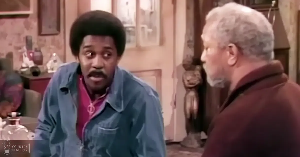 A middle-aged man and an older male seated indoors, wearing denim jackets over red shirts and maroon jackets respectively. The setting appears to be a dining room or kitchen with wooden furniture and bottles in the background.
