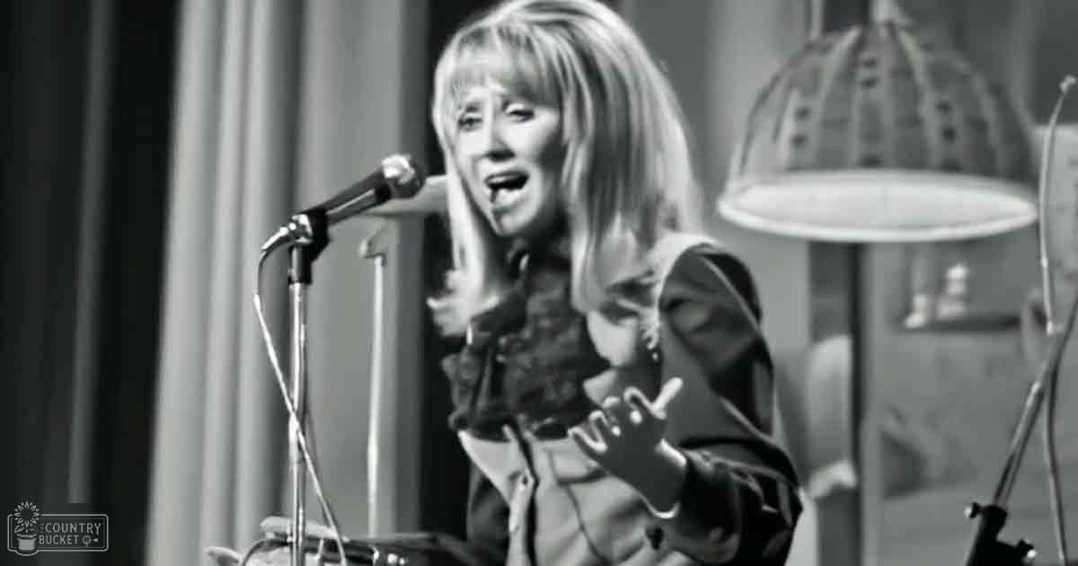 Middle-aged female singing into a microphone stand in what appears to be an indoor recording studio with curtains as the backdrop.