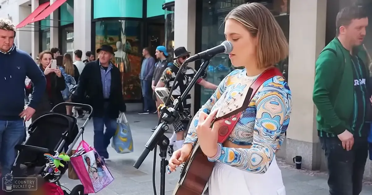 A young adult female street performer plays guitar on an urban city street, surrounded by passersby pushing strollers or holding shopping bags.