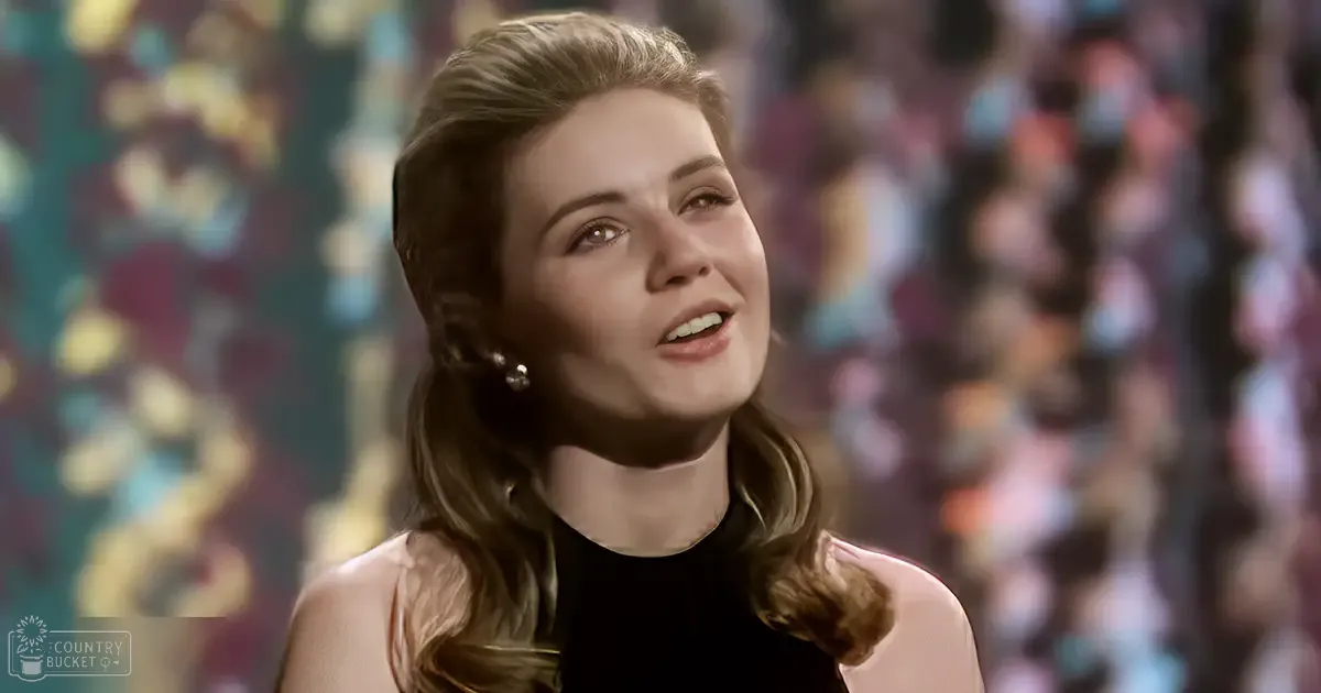 A young adult female wearing a sleeveless black top in an indoor performance setting with a crowd of spectators in the background, illuminated by artificial evening light.