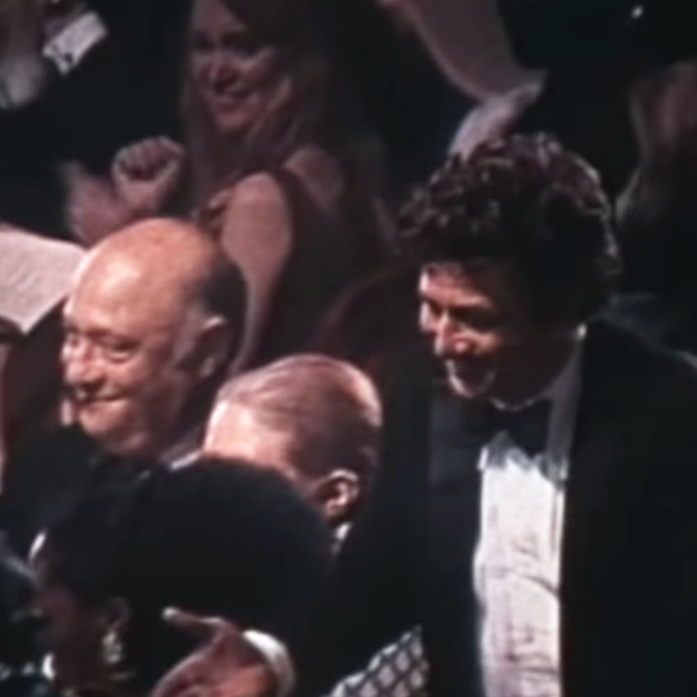 A group of adults at an indoor formal event, possibly awards ceremony; older adult male in dark suit with white shirt and black bow tie, middle-aged female in sleeveless dress or top, both from mid-20th century.