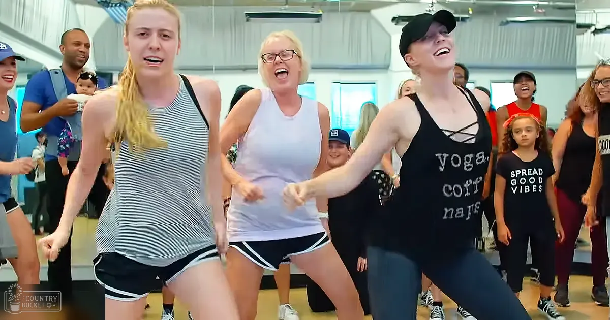 A group fitness class in a well-lit indoor gym setting, with young adults and middle-aged women wearing athletic attire.