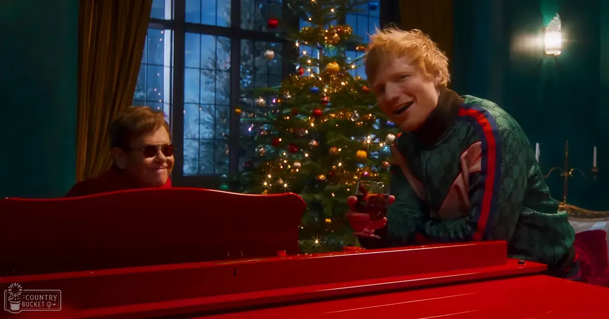 Two adults in a festive indoor Christmas setting with a large red piano, decorated tree, and snowy night background.