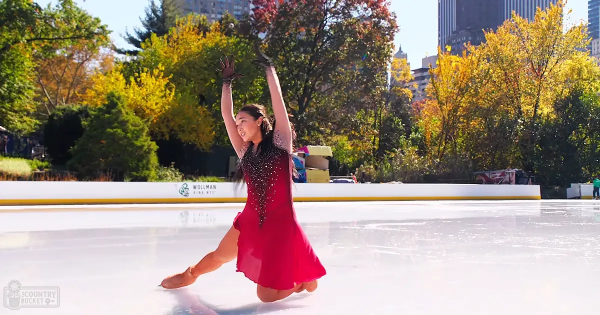 A young adult female figure skating at Wollman Rink in Central Park, New York City during fall with autumn foliage and clear blue skies.