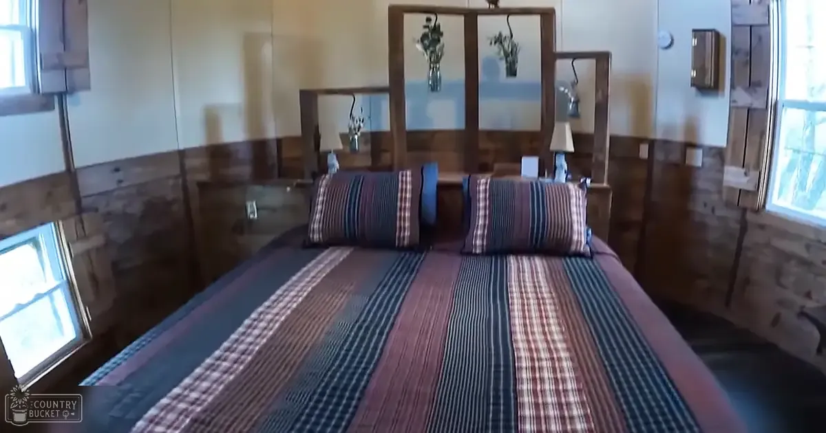 A bedroom scene with wood paneling on walls, a striped patterned bedspread in shades of blue, red, white, and black, two side tables with lamps, and natural light streaming through windows. The setting suggests winter or early spring.