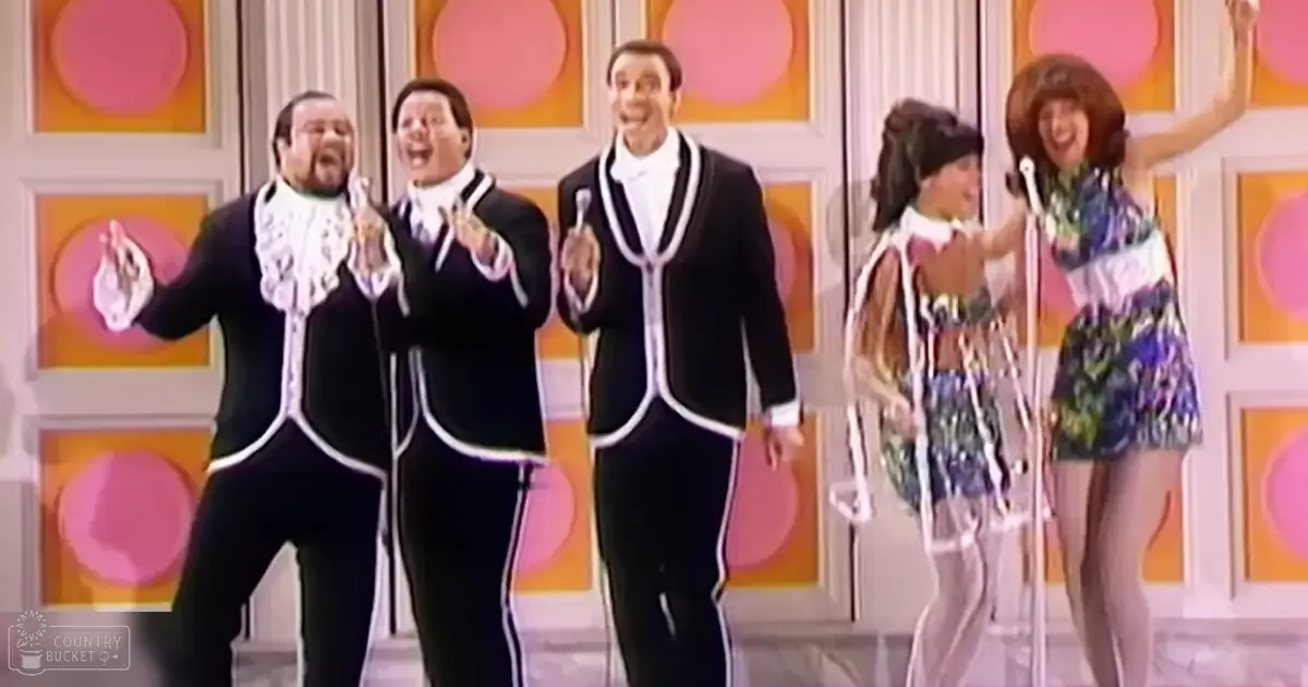 A group of performers in a mid-20th century studio setting, dressed in formal black tuxedos and a floral dress, against a vibrant wall with pink circles on an orange-yellow backdrop.