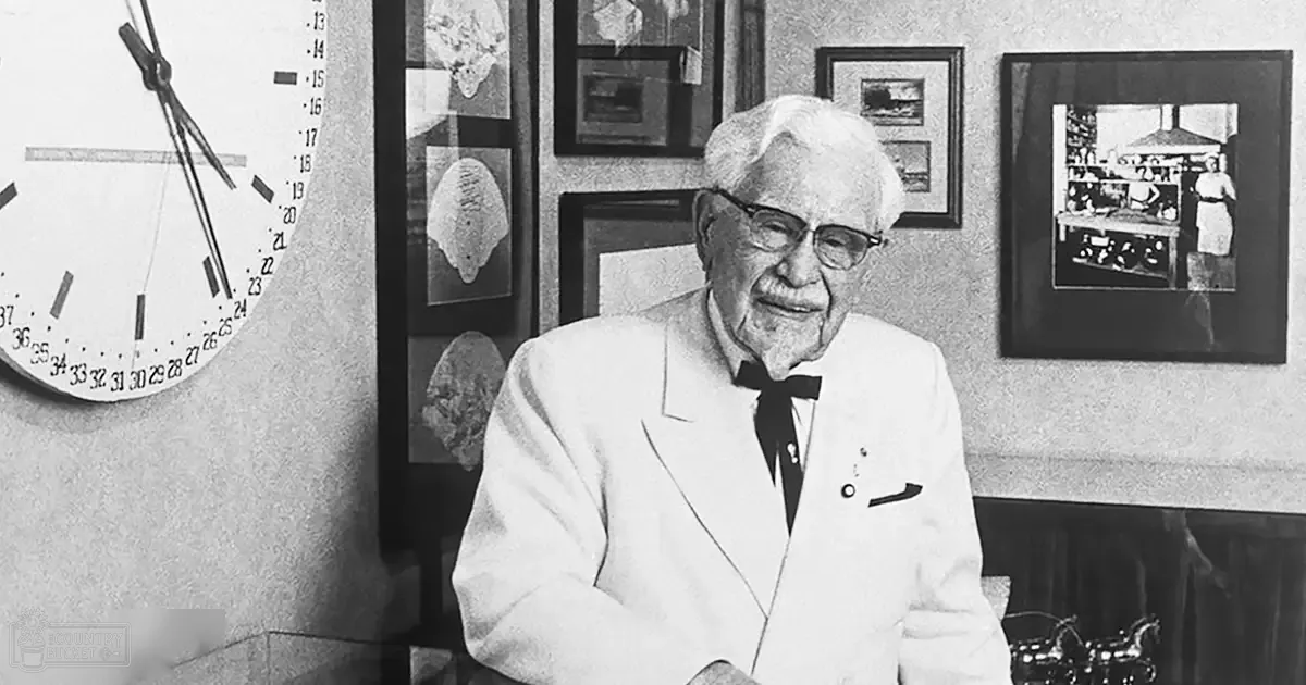 Colonel Sanders stands in his office, wearing a white suit jacket and bow tie.