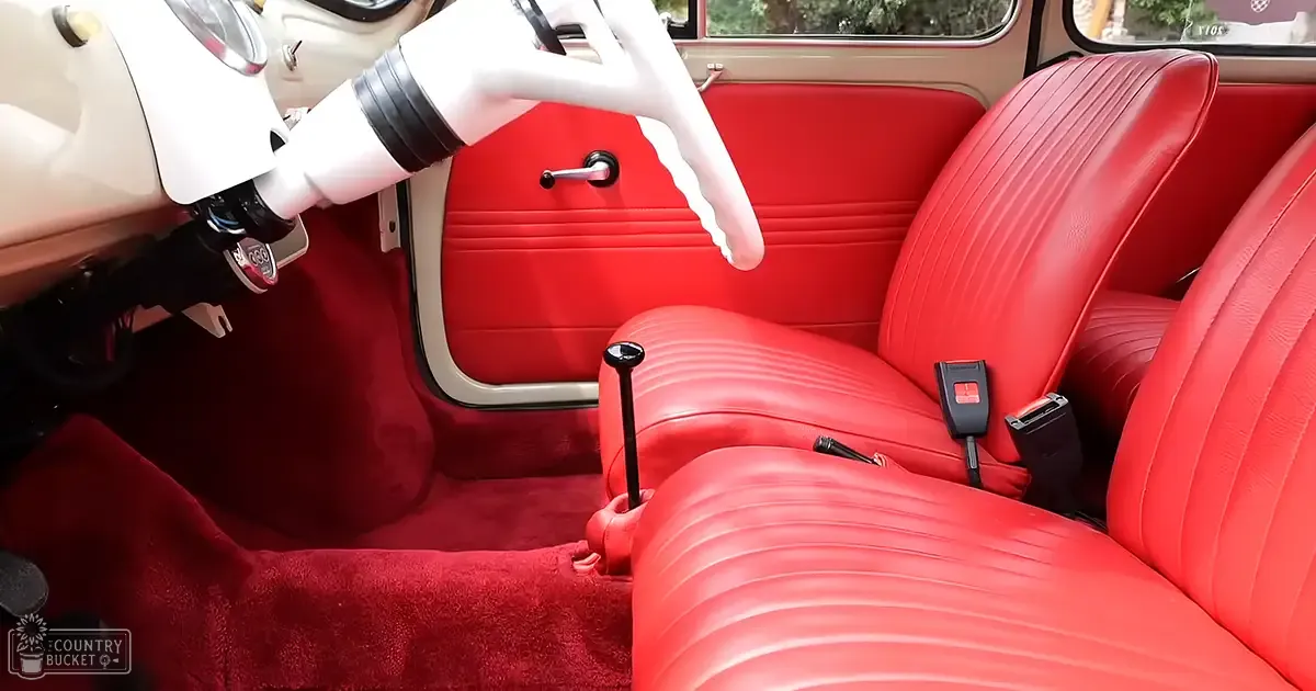Vintage car interior with red leather seats, white steering wheel, black gear shift in 1960s or early 1970s design; trees and natural light visible through windows outside.