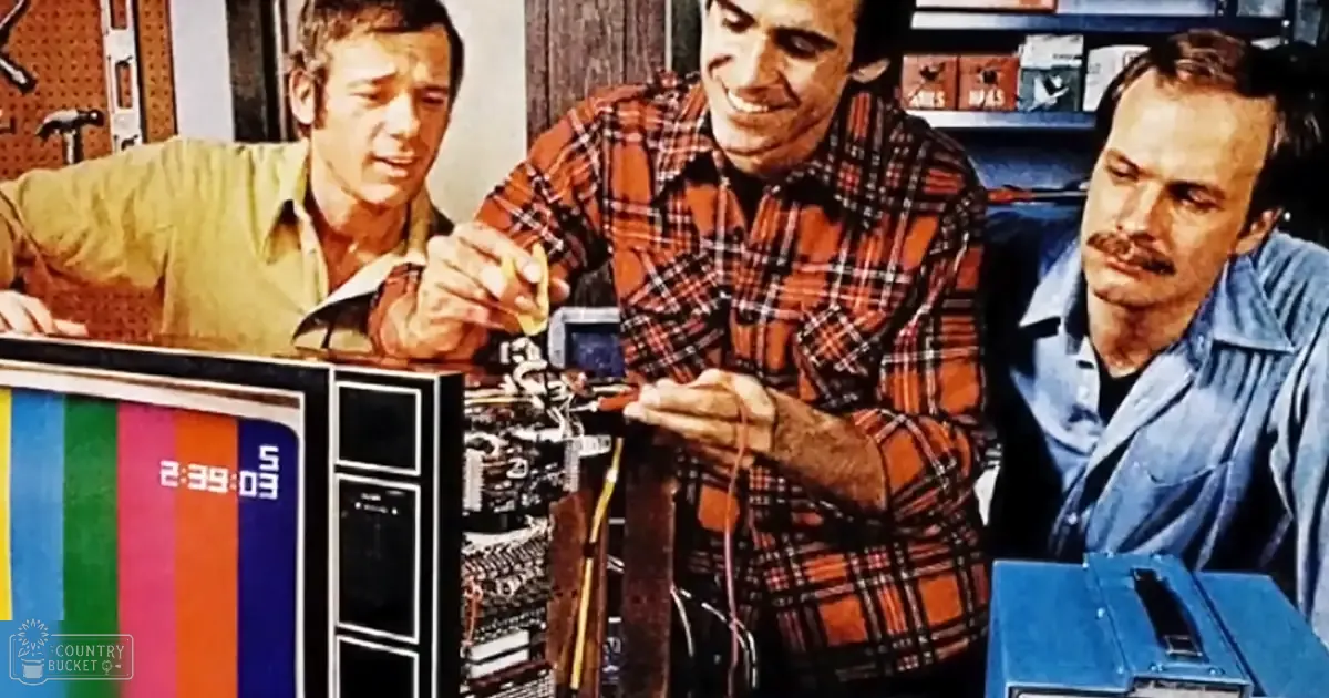 An adult male singer in a light green button-up shirt works alongside another middle-aged man holding an electronic component, with a third adult male in a blue button-up shirt observing nearby tools and electronics on a cluttered workbench indoors.