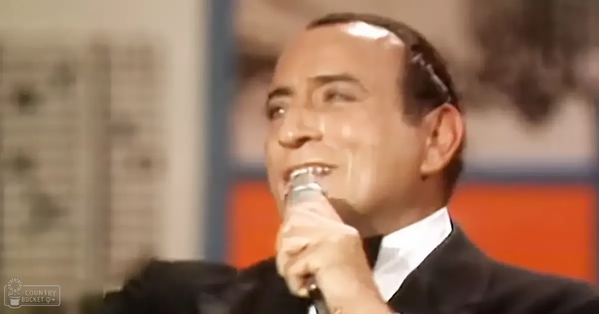 An adult male singer wearing a black suit jacket and white shirt, holding a microphone against a blurred studio backdrop with orange elements.