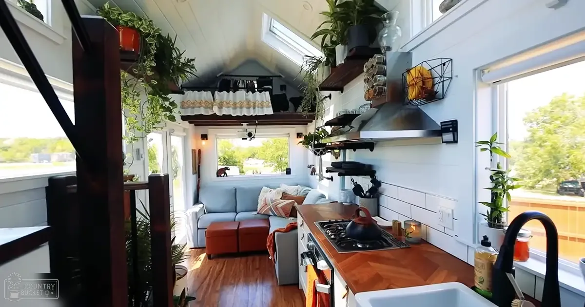 A cozy, well-decorated tiny house interior with a modern kitchen, open shelving, potted plants, and a light blue sofa in a residential setting during the daytime.