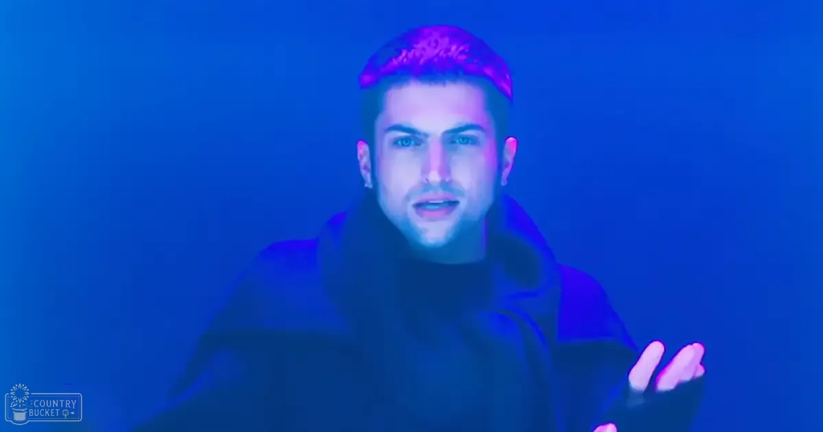 Young adult male wearing a dark-colored loose-fitting garment with a high collar indoors against a solid blue background, illuminated by blue light.