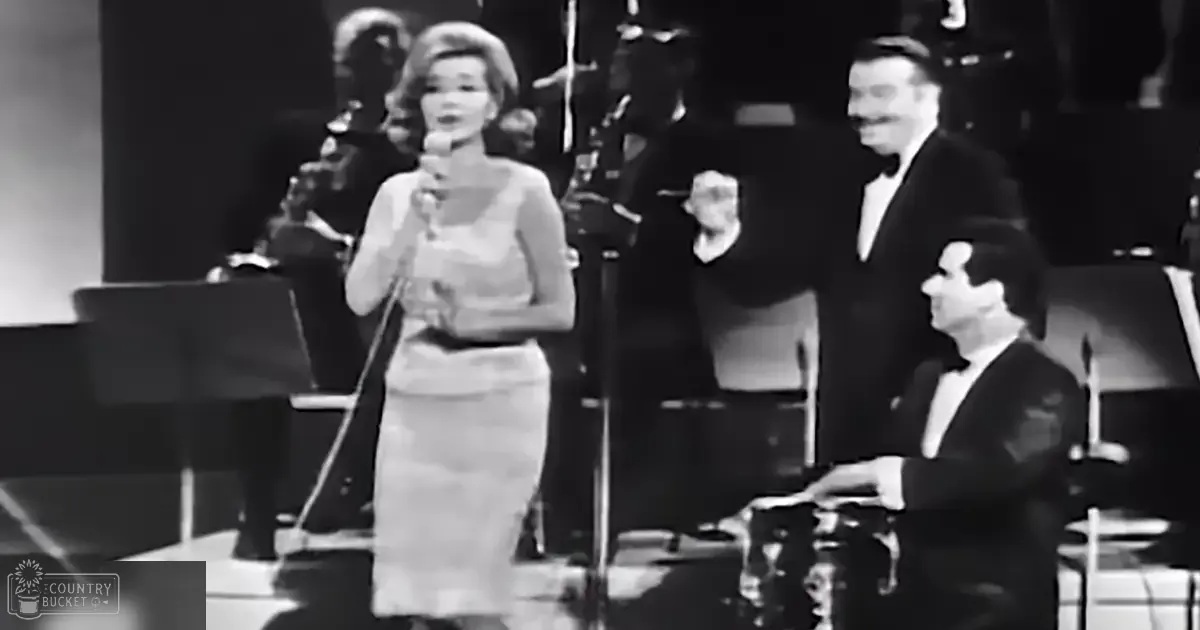 A woman singing into a microphone on stage before an orchestra; dressed in a light-colored knee-length dress, surrounded by formally dressed men in dark suits with white shirts and bow ties.