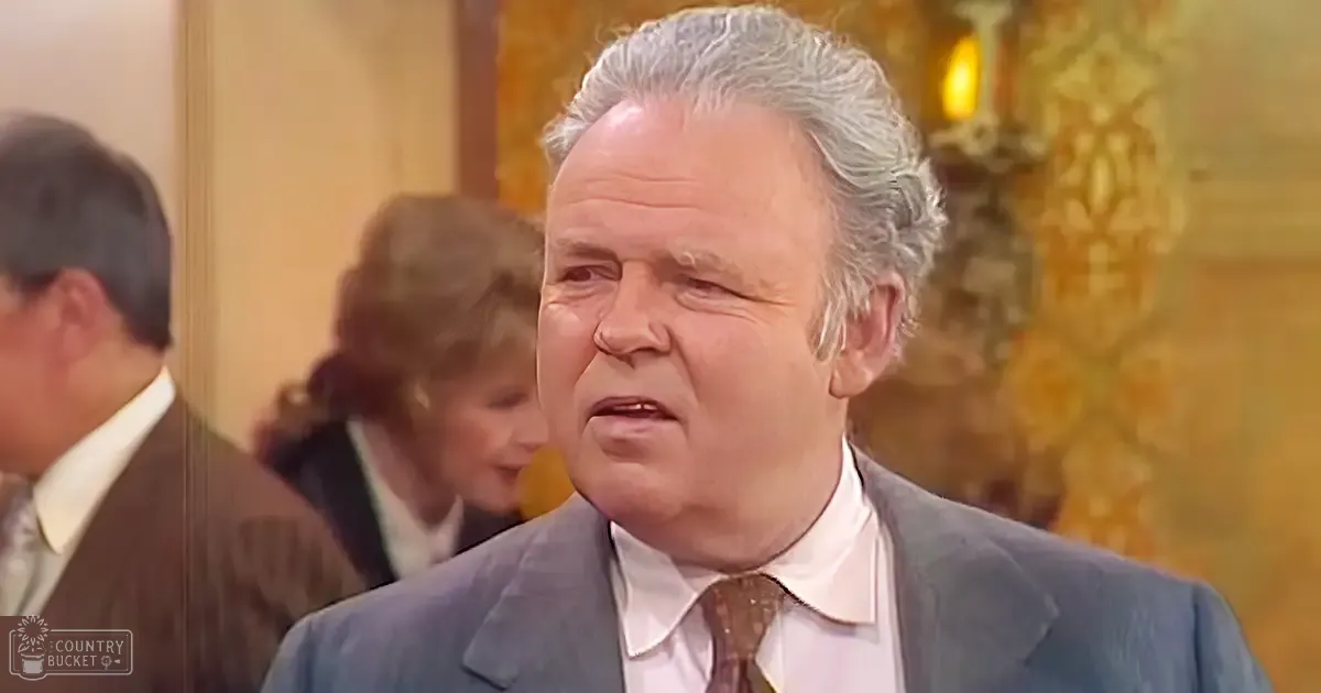Older adult male wearing a gray suit jacket, white shirt with brown and beige striped tie, indoors with ornate gold-patterned wallpaper, possibly an office or formal event space.