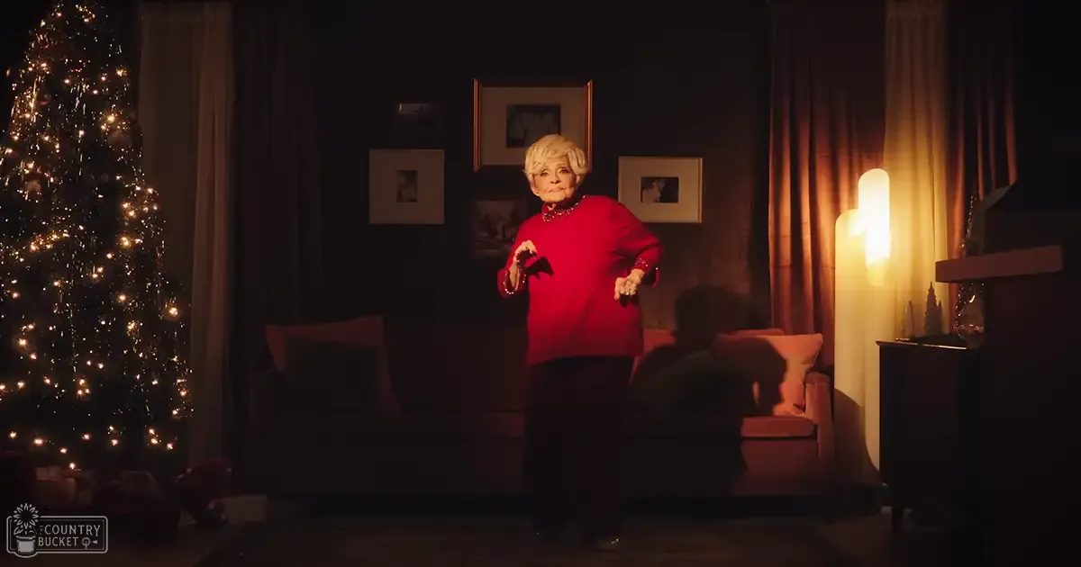 An older adult female wearing a red sweater and dark pants in a living room decorated for Christmas.