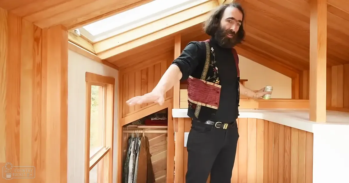 An adult male wearing a black shirt with floral embroidery and a red vest with gold trim, standing in an interior setting with wooden walls and ceiling.