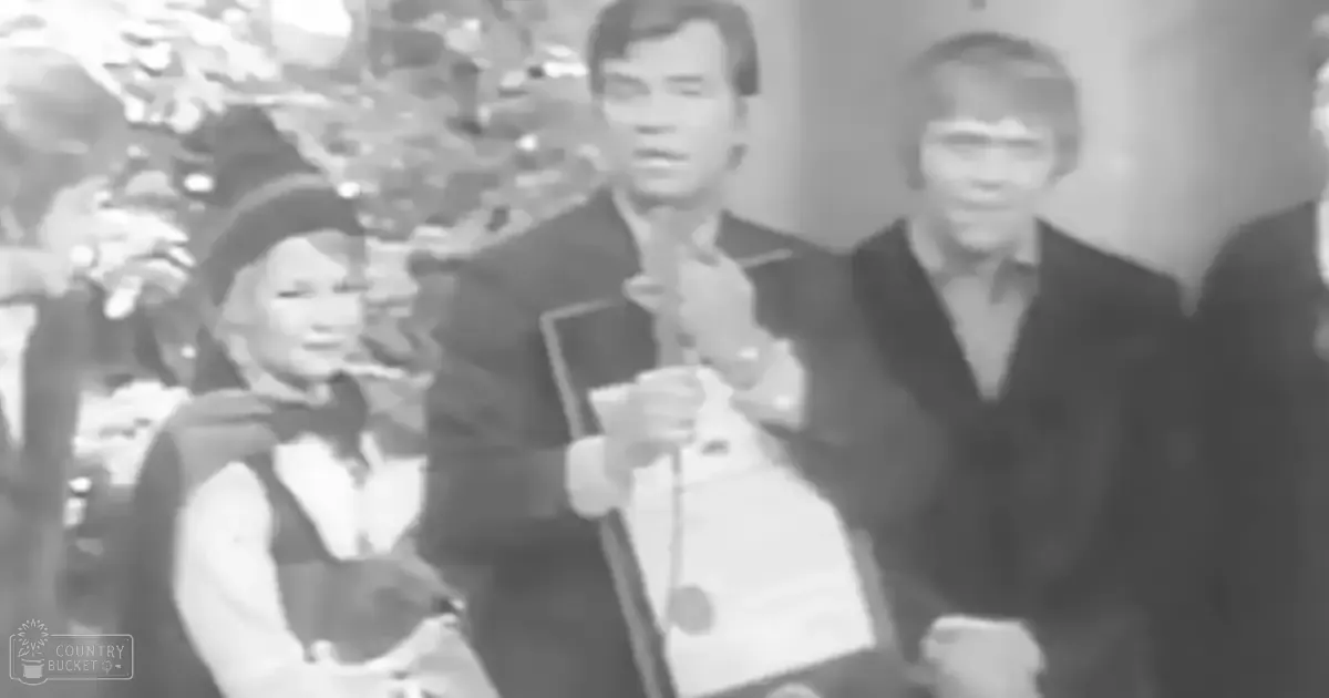 Three individuals dressed in mid-20th century attire gather around a decorated Christmas tree indoors under artificial lighting.