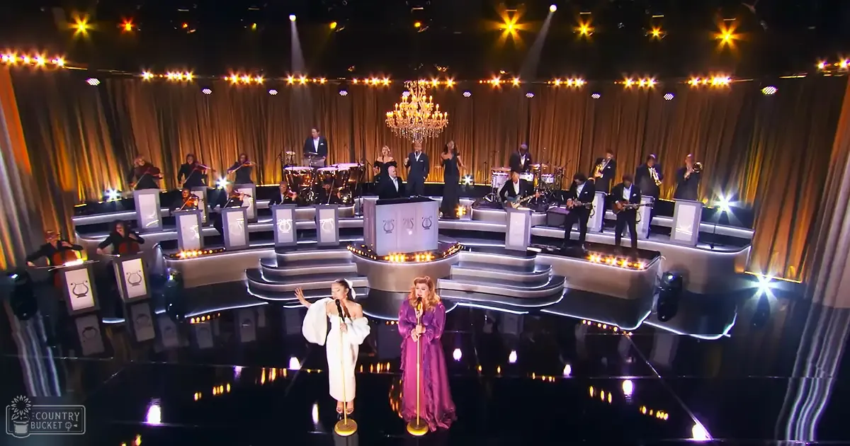 Musical performance scene in an indoor theater setting, with two young adult female singers in white dresses on stage, accompanied by musicians playing various instruments under warm artificial light.