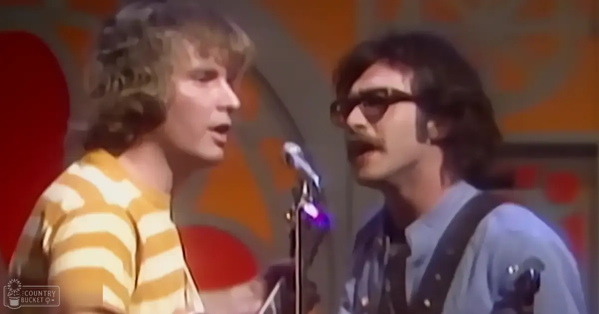 A young adult male singer wears a yellow and white horizontally striped shirt in an indoor performance setting with vibrant orange and red geometric patterns on the wall, suggesting a 1970s or early 1980s stage design.
