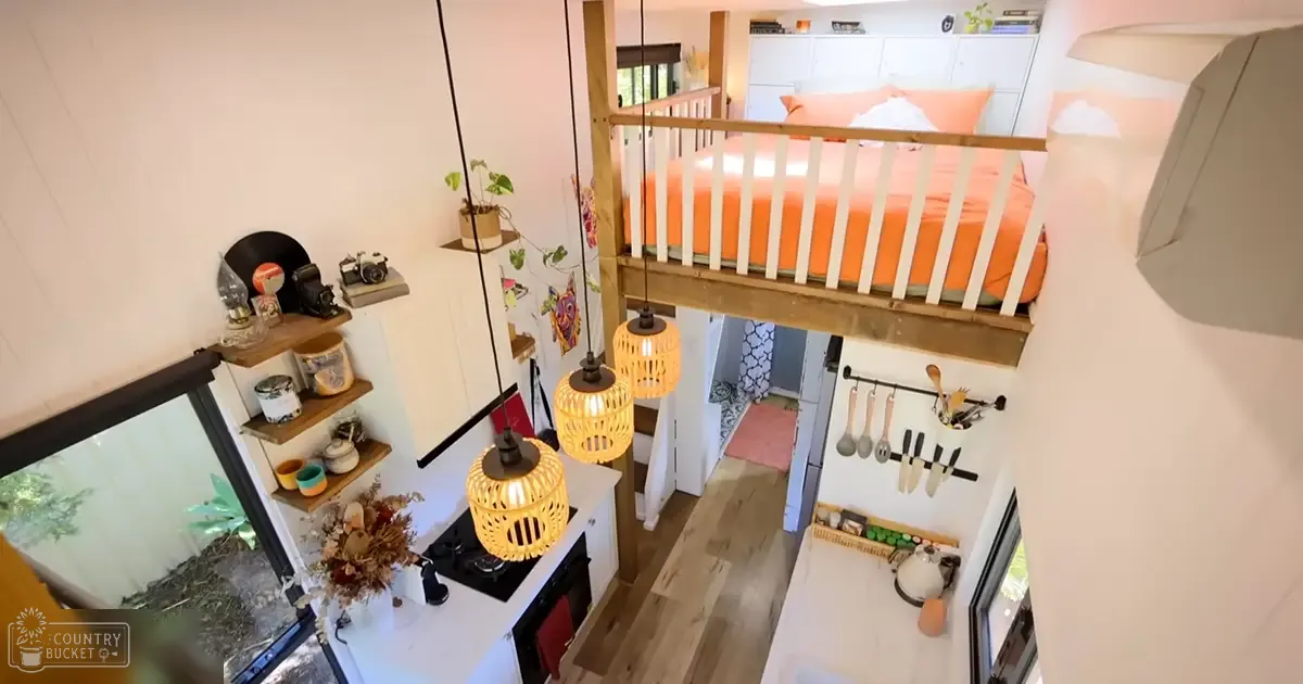 Cozy living space with loft bed, wooden shelves holding a record player and plants, small kitchenette, hanging pendant lights, window showing greenery outside; daytime.