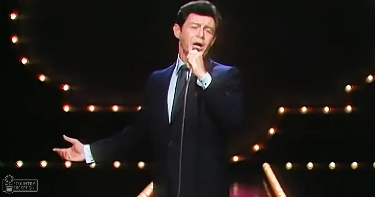 An adult male singer wearing a dark, well-fitted mid-20th century suit jacket and white shirt on stage with a microphone stand and circular string lights in the background.