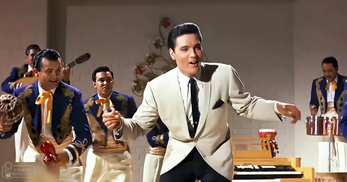 A male adult singer in a white suit jacket with black lapels, surrounded by other performers holding tambourines and maracas on a film set.