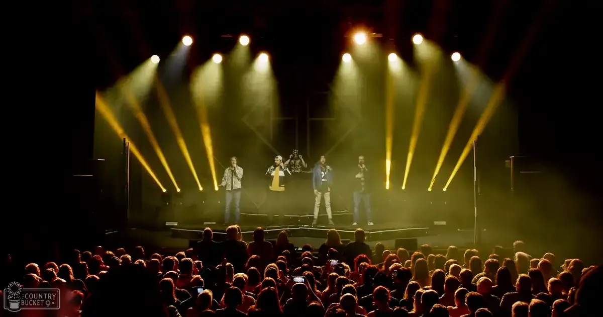 A group of adult male performers on a brightly lit stage, wearing casual attire against an indoor concert setting with yellow stage lights and dark audience seating.