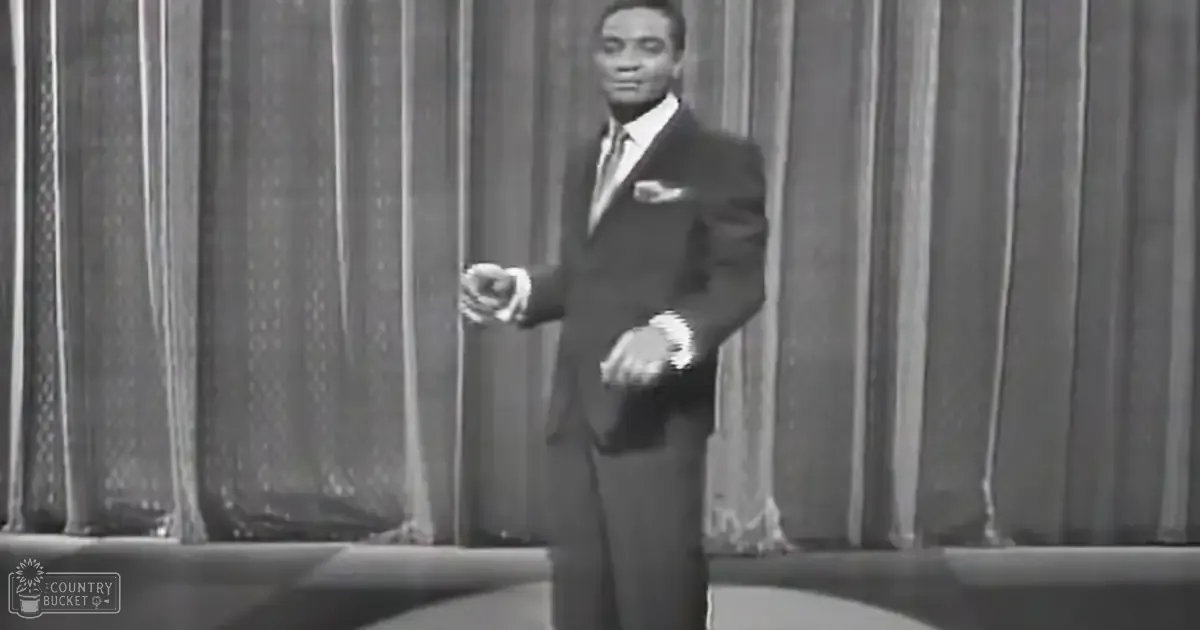 An adult male performer in a dark suit with a white shirt and tie stands on stage against a curtain backdrop under even lighting.