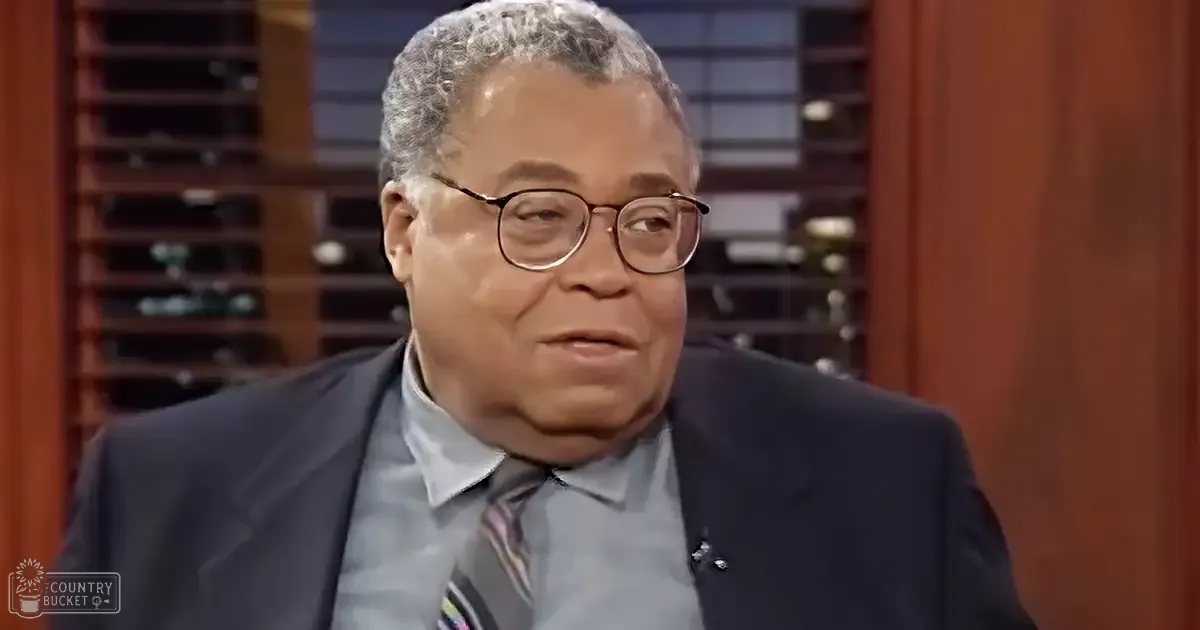 An older adult male in a studio setting wearing a dark suit jacket over a light gray dress shirt with a striped tie with shades of blue, pink, and white. The background includes wooden shelves and wine bottles.