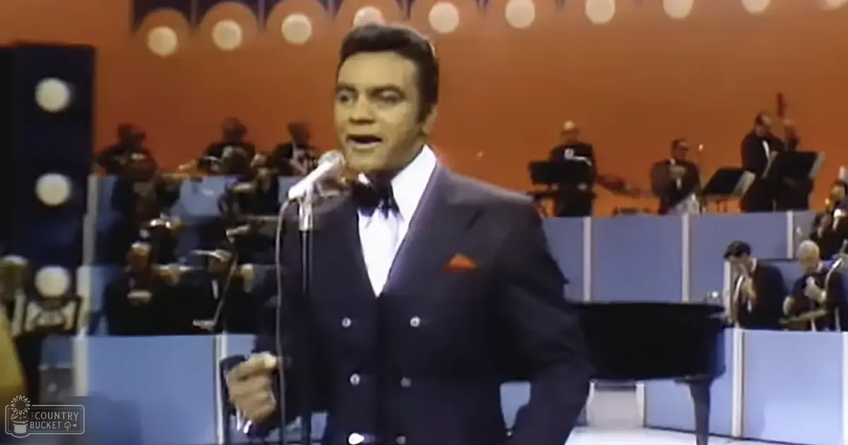 An adult male singer in a dark suit with white shirt and tie, holding a microphone, surrounded by musicians playing instruments in a well-lit studio setting with a grand piano and circular stage lights.