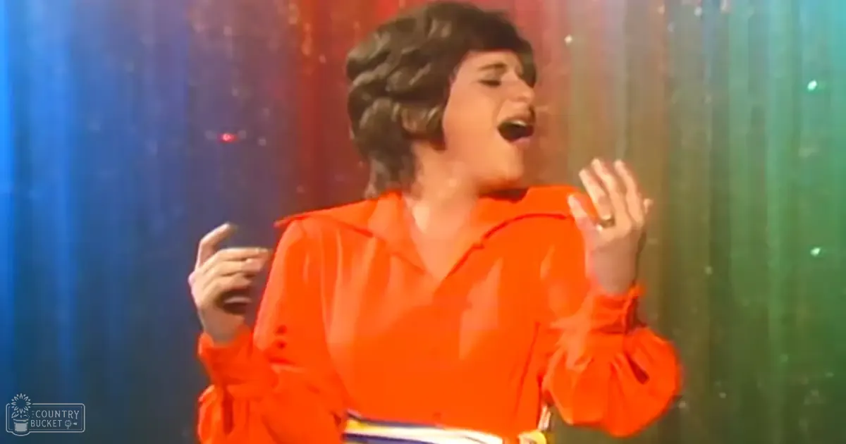 An adult female performer in a loose-fitting, bright orange blouse with a simple neckline and smooth fabric on an indoor stage with colorful lighting.