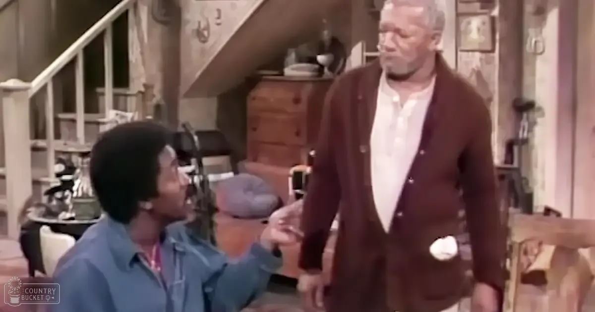 Young adult male wearing a blue button-up shirt with red collar, seated; older adult male in brown cardigan and white collared shirt standing indoors near staircase, wooden chest of drawers, globe, vases, and other household items.