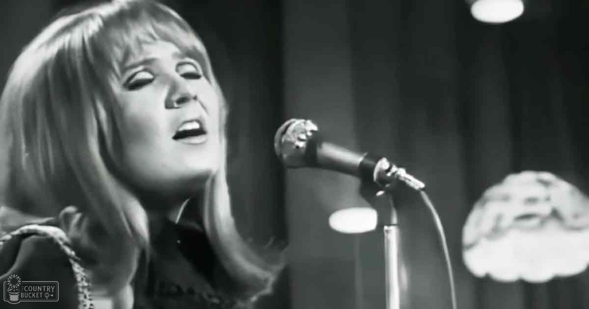 A young adult female with shoulder-length hair performing on stage in a dark-colored top with high neckline and long sleeves, set against curtains and stage lighting.