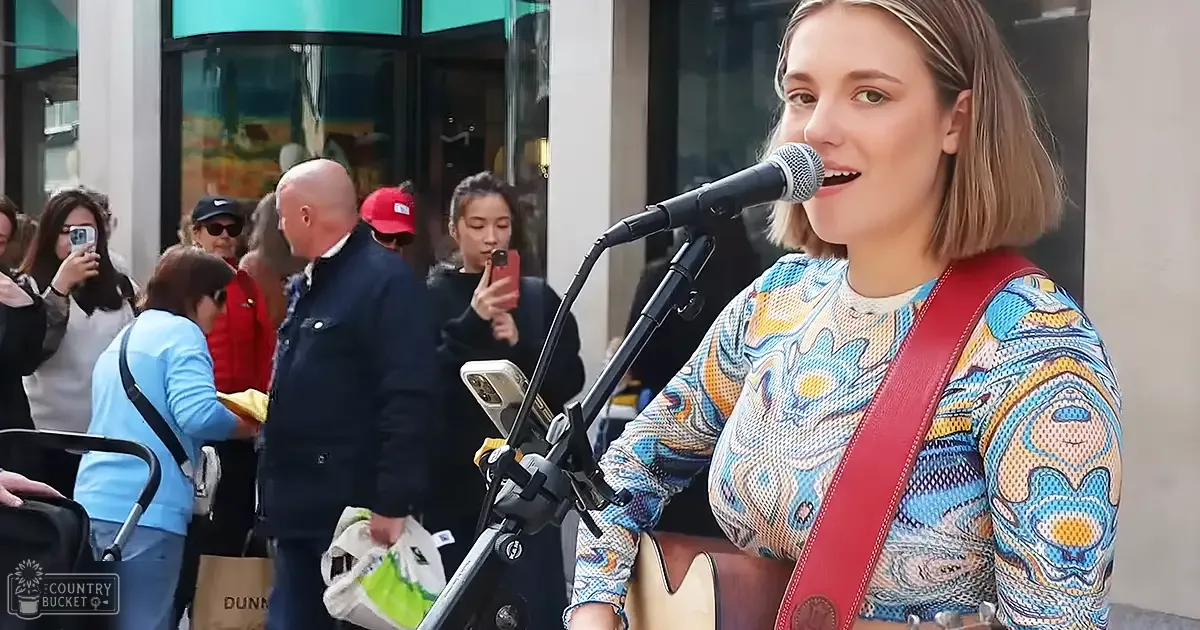 A young adult woman sings outdoors, wearing a colorful long-sleeved shirt with red guitar strap; an adult male holds shopping bags in the background.