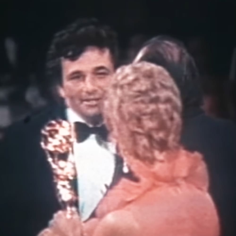 Adult male holding a golden Oscar statue at the Academy Awards ceremony in Hollywood, wearing a black tuxedo with white shirt and black bow tie.