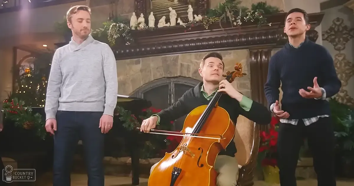 Adult males playing a cello in a living room decorated for Christmas with green sweaters, a fireplace adorned with religious figurines, and holiday garlands.