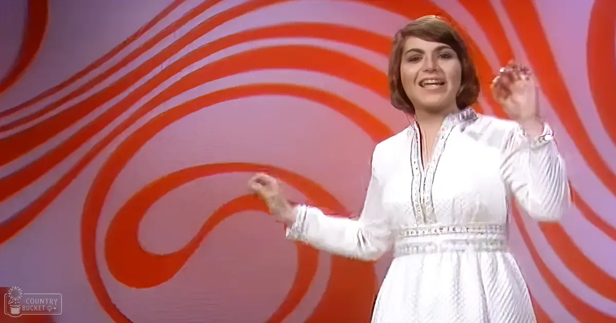 A young adult female wearing a white, flowy, high-necked dress with intricate embroidery, in an indoor setting with swirling red and white patterns behind her.