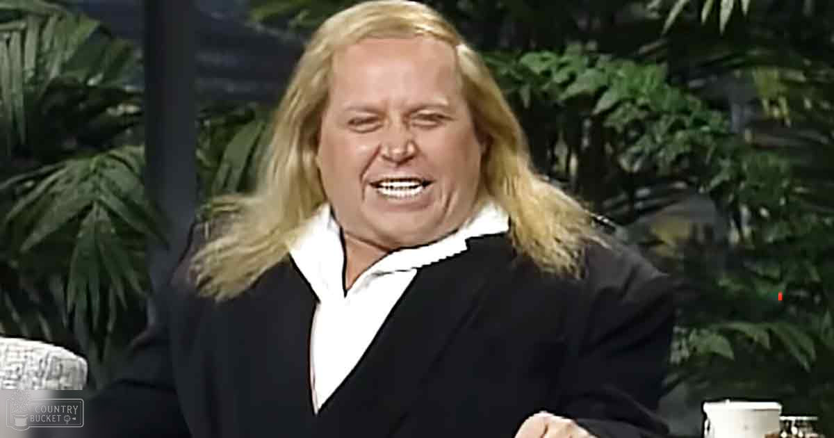 Middle-aged adult male wearing a black blazer with white shirt, seated indoors possibly on a talk show or interview set with patterned furniture and green foliage in the background.
