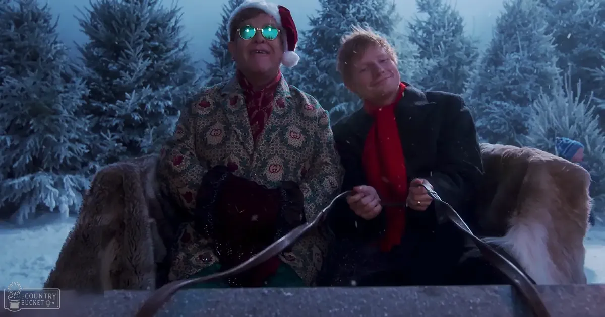 Two middle-aged men in a snowy outdoor setting, possibly during Christmas time, wearing festive clothing and holding what appears to be a piece of paper or ribbon.