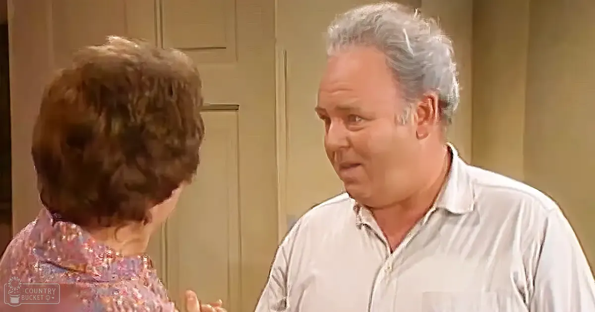 A middle-aged man with gray hair in a light-colored, striped collared shirt stands indoors alongside an older adult woman with short brown hair styled in an updo wearing a patterned blouse or dress with small floral designs. The setting is simple and appears to be daytime with even lighting.