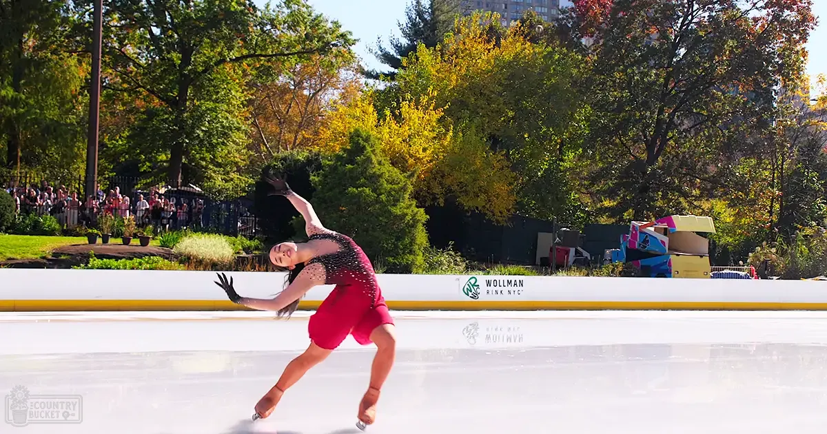 A young adult female figure skater in a red, embellished figure skating costume at Wollman Rink NYC on an autumn day with trees displaying green and yellow leaves.
