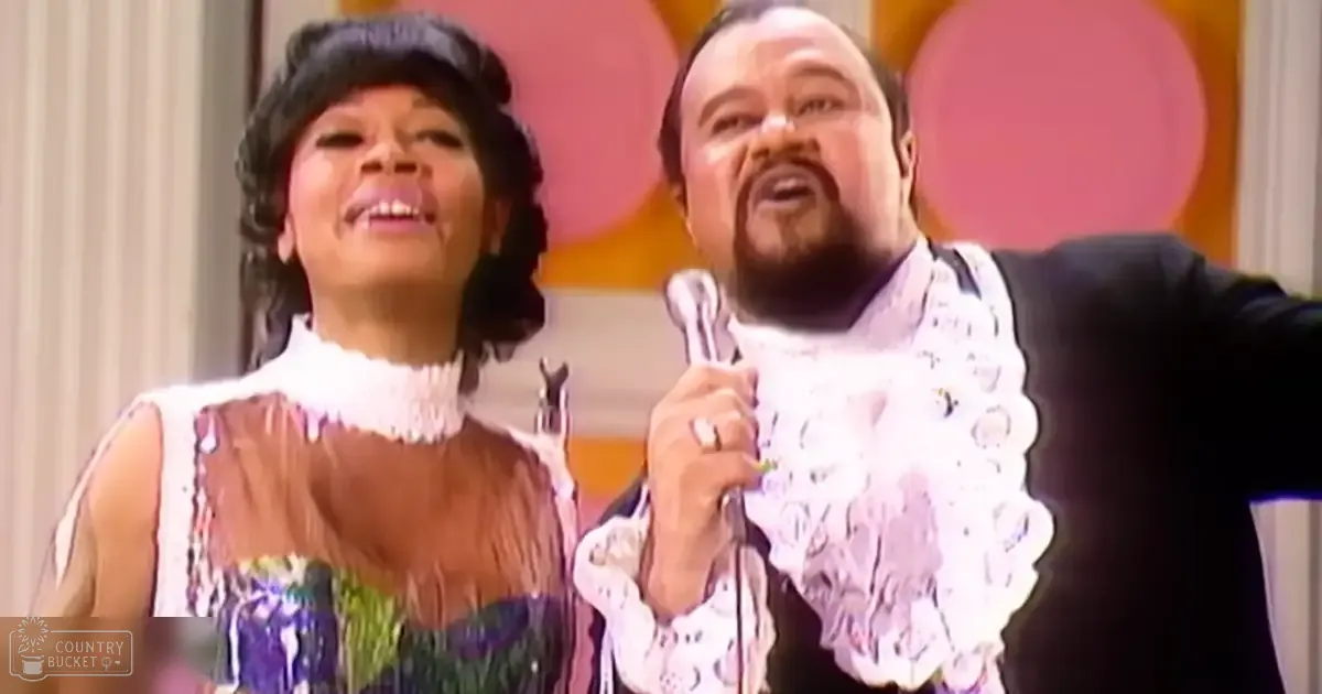 Musical performance scene with an adult female singer wearing a sleeveless dress and an adult male performer in a black suit, set on an indoor stage under warm artificial lighting.
