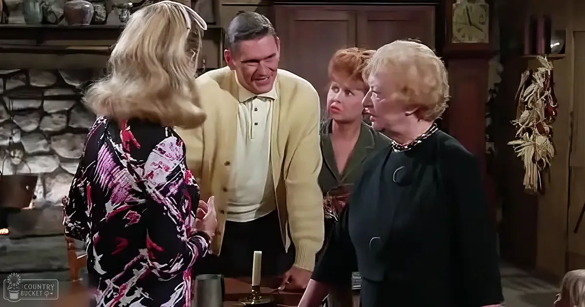 A cozy indoor scene with a middle-aged woman in a floral dress and an older adult female in a dark green wool dress gathered around a wooden table. Corn husks hang on the wall, and a stone fireplace with hanging pots and pans is visible behind them.