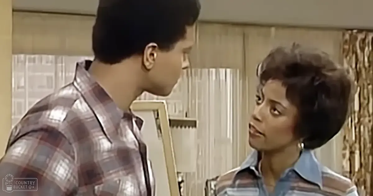 Young adult male in plaid shirt with reds, blues, and whites, and young adult female in striped blue, white, possibly pink or purple shirt with hoop earrings. Setting is a living room with curtains and chair.