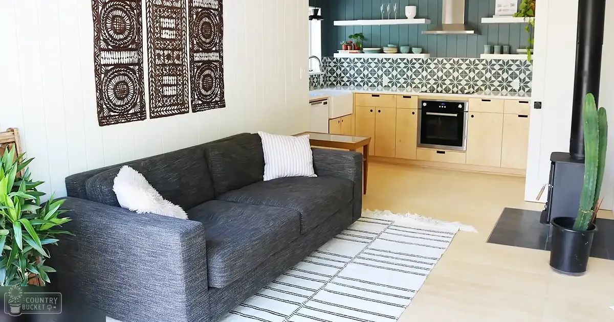 An indoor living room scene with a dark gray sectional sofa, white pillows, black and white striped rug, wooden coffee table, light brown cabinets, black stove/oven unit, decorative wall hangings, and green plants. The setting appears to be indoors in a home during daytime.