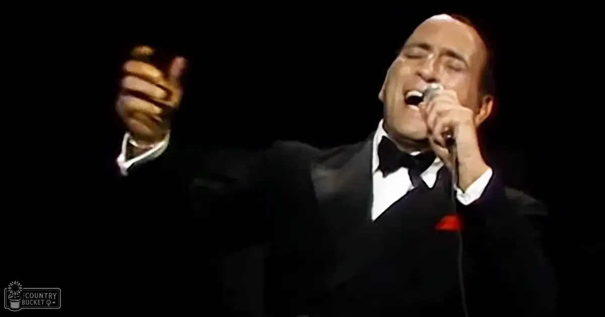 A middle-aged male performer in a black tuxedo holding a microphone, possibly singing on stage.