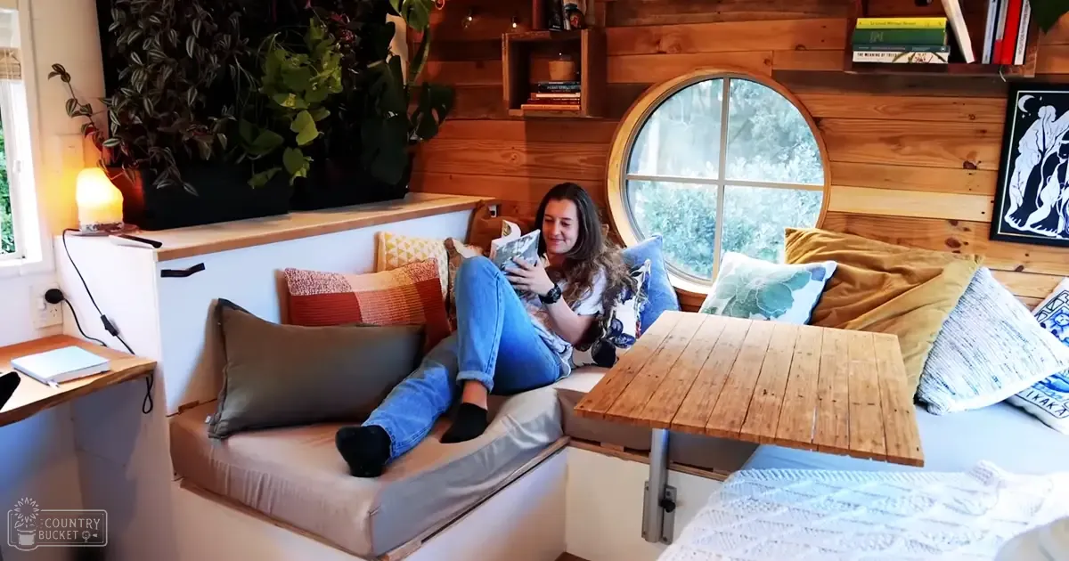 A young adult female in a cozy indoor setting, possibly a cabin or tiny home. She is wearing jeans and a white t-shirt with a floral pattern, accessorized with a wristwatch, surrounded by rustic furniture and decor.