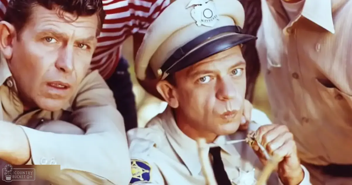 A town square scene from 'The Andy Griffith Show' with individuals dressed in period attire, including a man in a white police uniform holding handcuffs, and a crowd of people in various clothing styles.